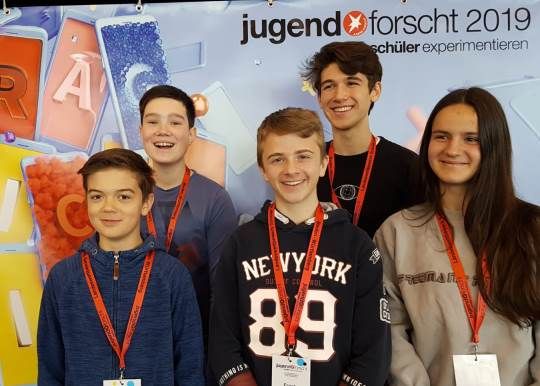 Fünf Jugendliche mit Teilnehmerausweisen stehen vor der Wand von Jugend forscht und Schüler experimentieren 2019