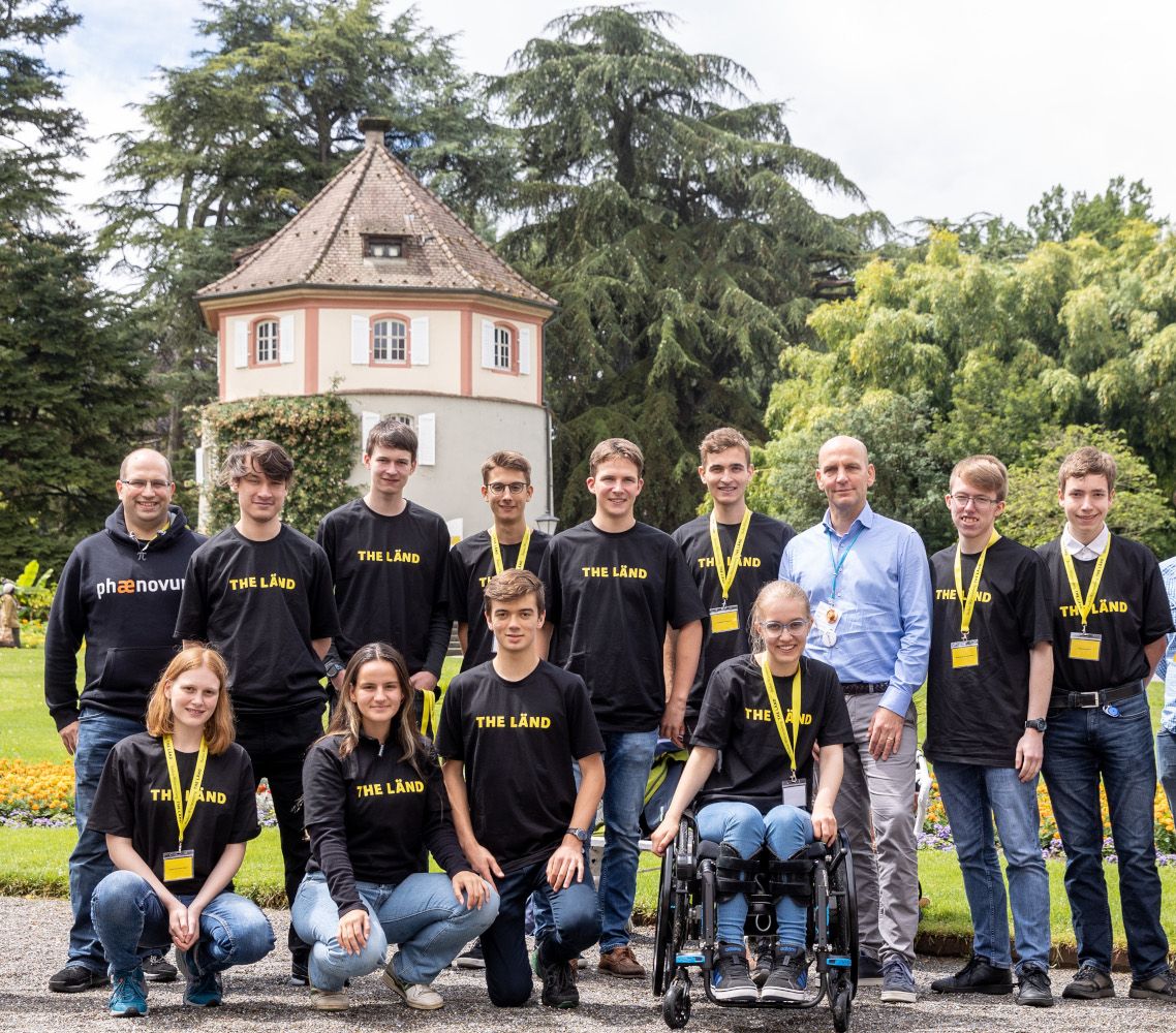 Gruppenfoto einer Jugendgruppe in schwarzen Shirts mit der Aufschrift „THE LÄND“ in einem Park vor einem kleinen Turmhaus