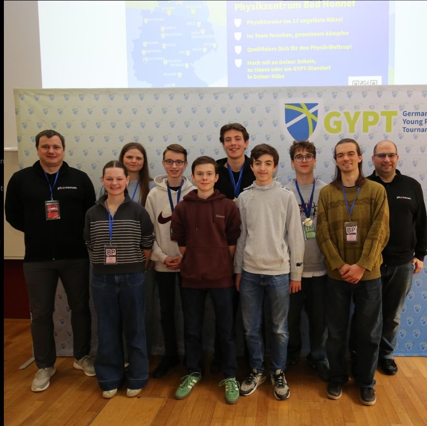 Das Team steht mit seinen Betreuern vor der Wand des German Young Physicists' Tournament, auf der Leinwand dahinter die Ankündigung des Physikzentrums Bad Honnef