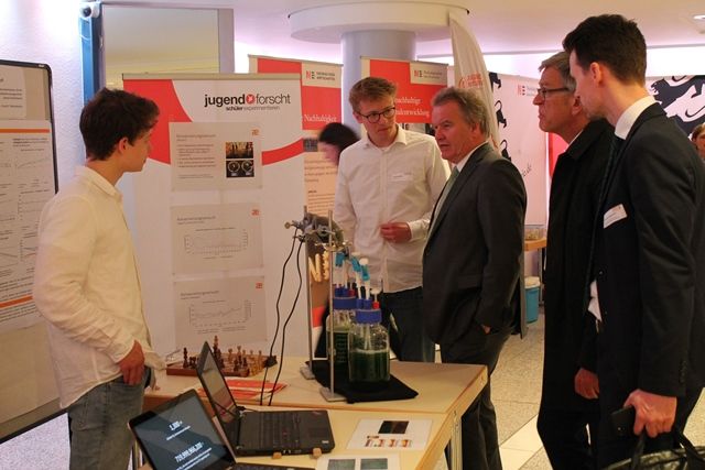 Ein Jungforscher erklärt Besuchern im Anzug seinen Versuchsaufbau mit grünen Algenkulturen am Jugend-forscht-Stand