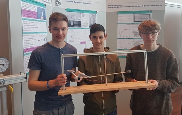 Drei Jungforscher halten ihren Versuchsaufbau zum Ionenwind vor ihre Posterwand
