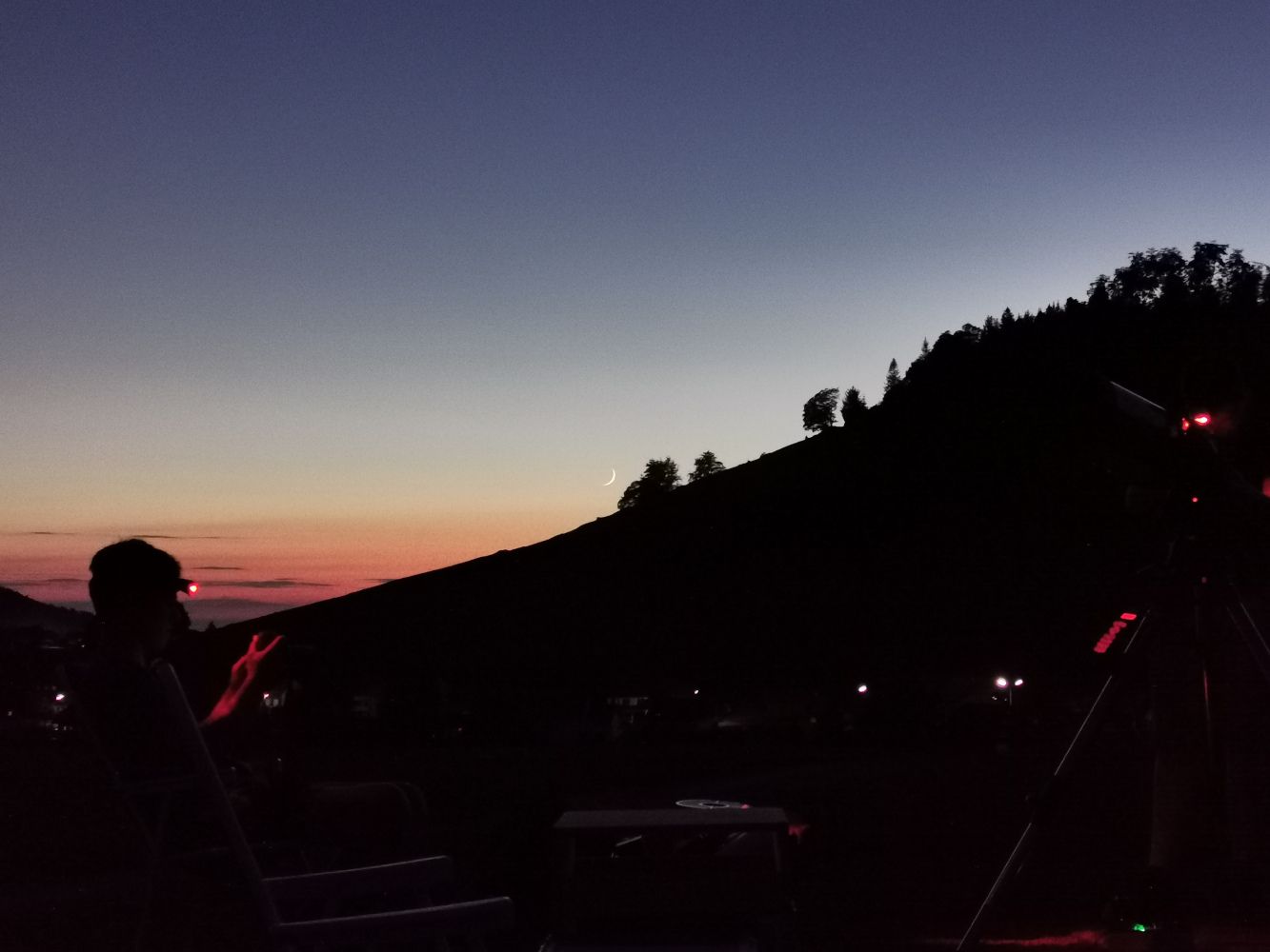 Schmale Mondsichel über dem Bergrücken in der Abenddämmerung, davor die Silhouetten von Beobachtenden und Teleskopen