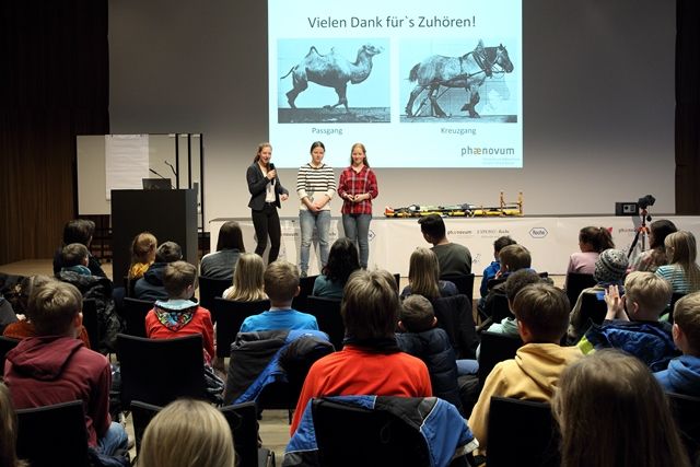 Drei Schülerinnen stehen vor einem vollen Saal auf der Bühne, auf der Leinwand die Schlussfolie ihres Vortrags über Passgang und Kreuzgang
